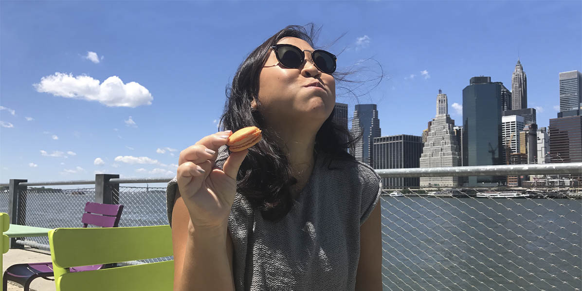 A dark-haired woman has a face of delight while eating a Peaches & Cream French macaron. Behind her is the New York skyline.
