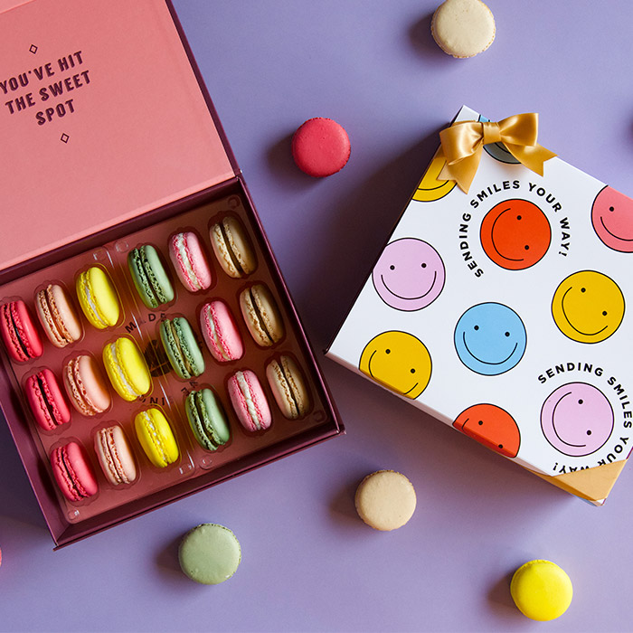 A purple background features a Woops! box of 18 French macarons with assorted flavors and a white sleeve with colored smiley faces with a golden ribbon. Some French macarons are lying around.