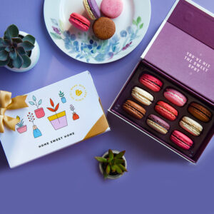 A purple background features a Woops! box of 9 French macarons with assorted flavors, a plate with 4 macarons, some green plants, and a white sleeve with pot figures at the center.