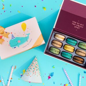 A blue background features a Woops! Box of 9 French macarons to the right, beside it a cream sleeve with a whale in the center and a golden ribbon. Surrounding them are some candles, confetti, and a party hat.