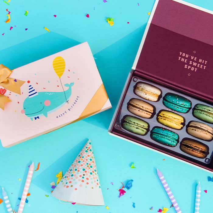 A blue background features a Woops! Box of 9 French macarons to the right, beside it a cream sleeve with a whale in the center and a golden ribbon. Surrounding them are some candles, confetti, and a party hat.