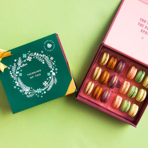 A green background features a Woops! box of 18 French macarons with assorted flavors, to its left is a dark green sleve with an ornament figure at the center and a golden ribbon.
