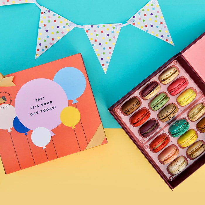 A blue and yellow background features a Woops! Box of 18 French macarons with assorted flavors and a red sleeve with colored balloons and a golden ribbon. At the top is a party decoration.