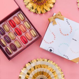 A pink background features a Woops! Box of 18 French macarons with a white sleeve with letters and a golden ribbon. At the top and bottom are some golden party decorations.