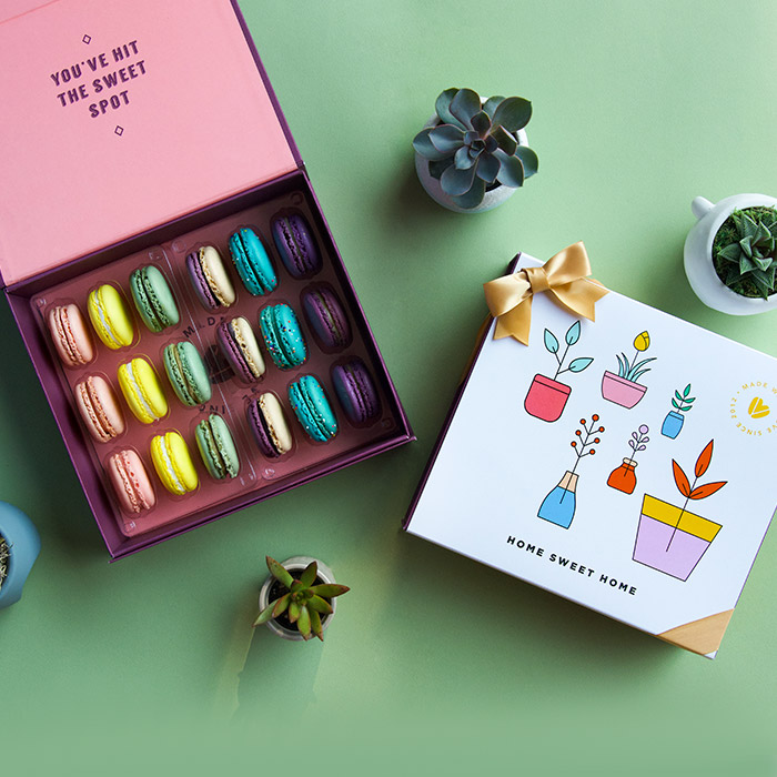 A green background features a Woops! box of 18 French macarons with assorted flavors, some green plants, and a white sleeve with pot figures at the center and a golden ribbon.