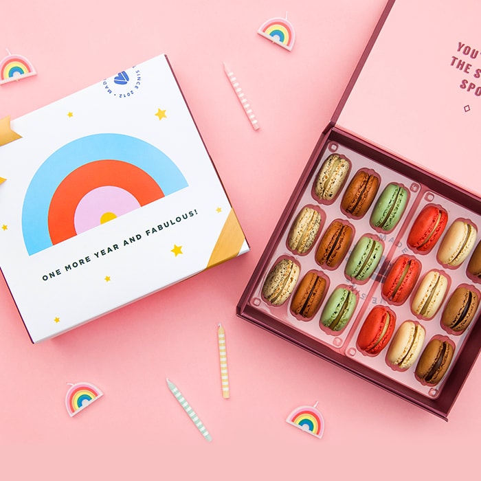 A pink background features a Woops! Box of 18 French macarons with assorted flavors, to its left is a white sleeve with a rainbow in the center and a golden ribbon. Surrounding them are some candles, party decorations, and tiny rainbows.