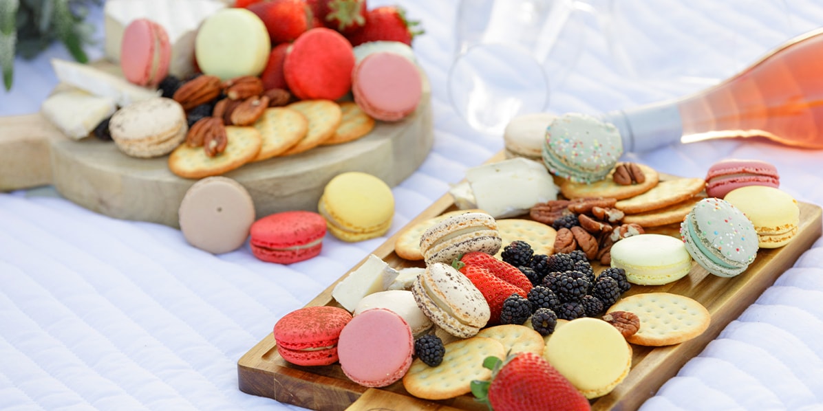 A white mantle features 2 wooden cheese tables full of fruits, crackers, and Frenchs macarons with assorted flavors, a bottle of wine and some glasses