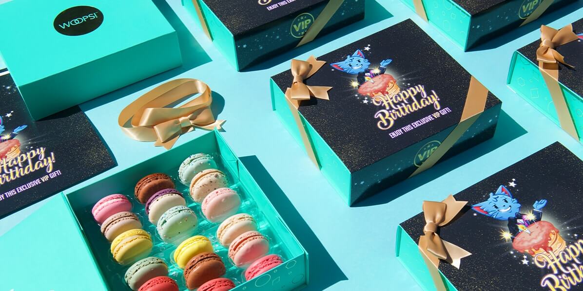 An aquamarine background features an open box of 18 French macarons with assorted flavors surround by customized Playtika Birthday boxes with golden ribbons.