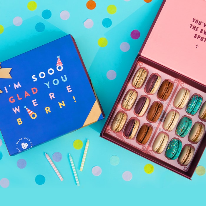 A blue background with colored dots features a Woops! Box of 18 French macarons with assorted flavors, to its left is a dark blue sleeve with colored letters and a golden ribbon. Surrounding them are several candles.