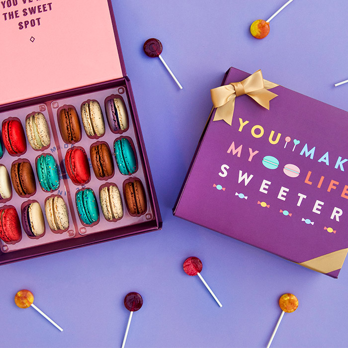 A purple background features a Woops! box of 18 French macarons with assorted flavors and a purple sleeve with colored letters at the center plus a golden ribbon. Some lollipops are lying around.