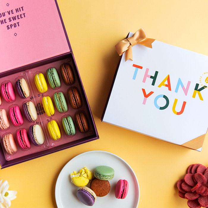 A yellow background features a Woops! box of 18 French macarons with assorted flavors, a white sleeve with a big colored "thank you" at the center and a golden ribbon, plus a plate with French macarons and some flowers.