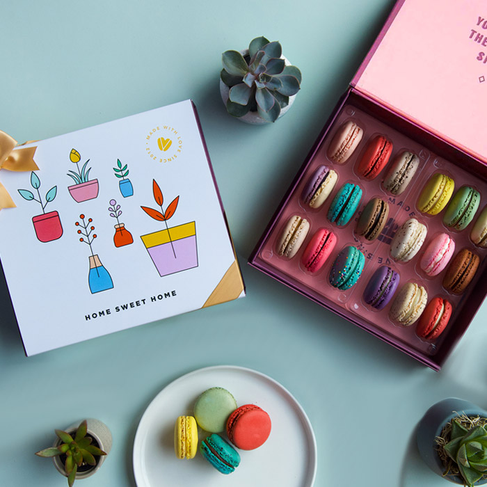 A green background features a Woops! box of 18 French macarons and a white sleeve with some pot drawings in the middle with a golden ribbon. Some green plants are lying around and at the bottom is a plate with 4 French macarons.