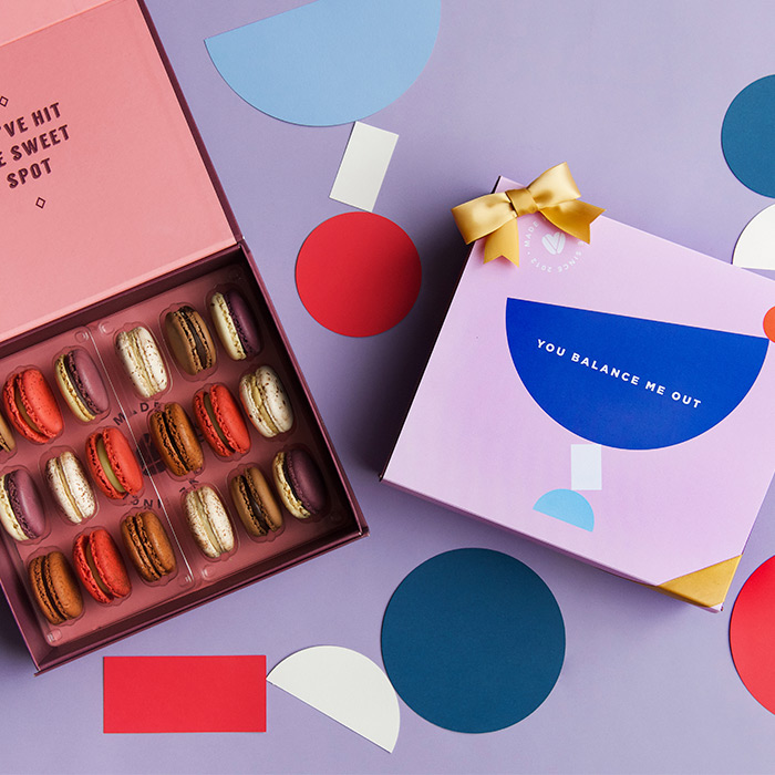 A purple background features a Woops! box of 18 French macarons with assorted flavors, to its right is a light purple sleeve with a blue figure at the center and a golden ribbon. Some cardboard geometrical figures are lying around.