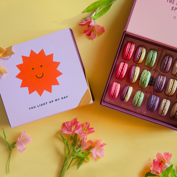 A yellow background features a Woops! box of 18 French macarons with assorted flavors and a light purple sleeve with a smiling sun in the middle plus a golden ribbon. Some pink flowers are lying around.