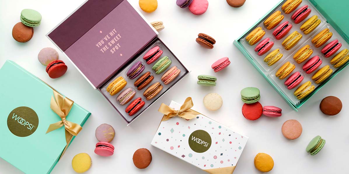 A white background features a Woops! Box of 18 French macarons with Strawberry and Lemon Tart flavors, a box of 9 French macarons with assorted flavors, a green closed Woops! Box and a white closed Woops! Box with golden ribbons. Lying around are assorted French macarons.