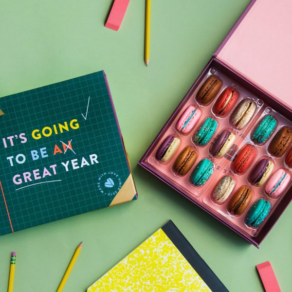 A box full of 18 assorted macarons has a macaron box with a green B2S sleeve to the left. Surrounding them are school supplies.
