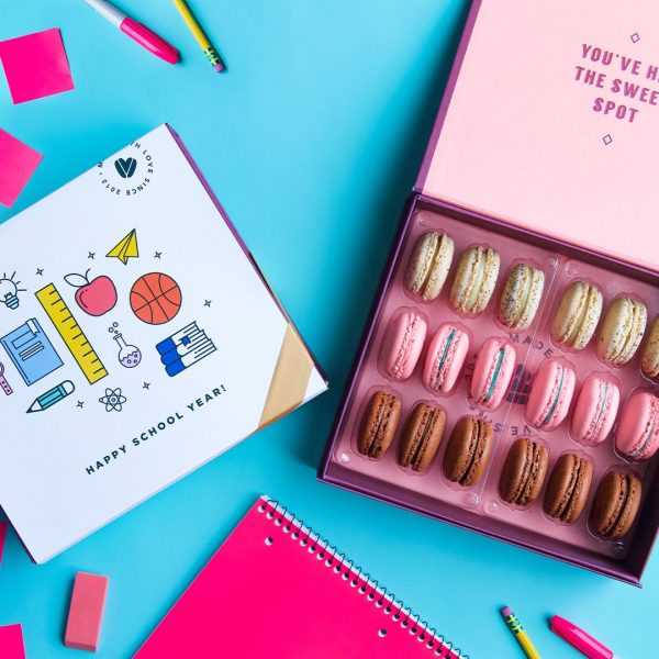 A box full of 18 assorted macarons has a macaron box with a white B2S sleeve to the left. Surrounding them are school supplies.