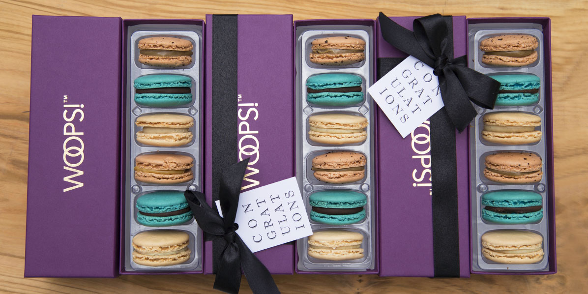 3 purple French macaron boxes with assorted flavors are on top of a wooden board.