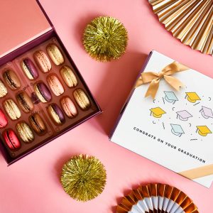 A box full of 18 assorted French macrons has a macaron box with a Graduation sleeve to the right. Surrounding them are some golden party decorations.