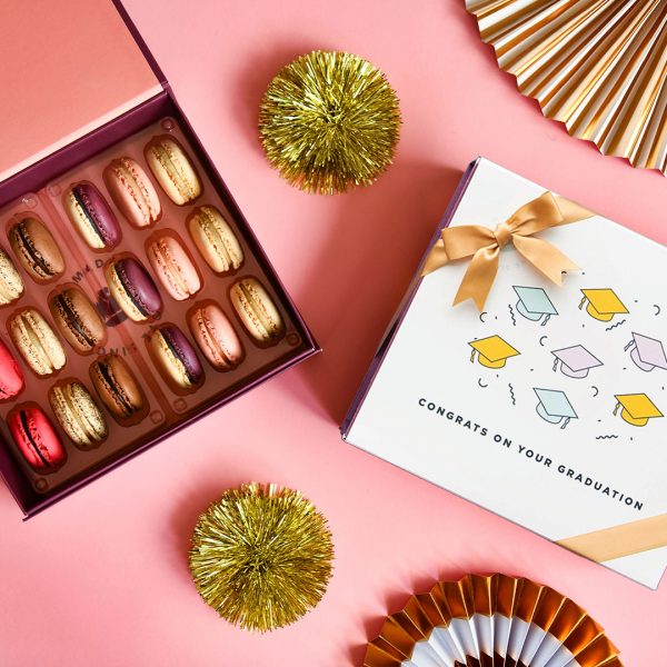 A box full of 18 assorted French macrons has a macaron box with a Graduation sleeve to the right. Surrounding them are some golden party decorations.