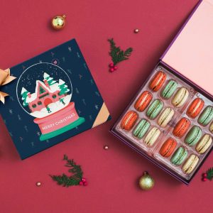 A box full of assorted macarons has a macaron box with a blue Holiday sleeve to the left.