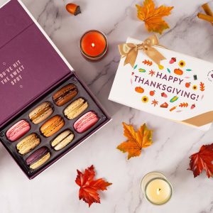 A box full of assorted macarons has a Woops! macaron box with a Thanksgiving sleeve to the right. Surrounding them are some pumpkins, acorns, and paper leaves.