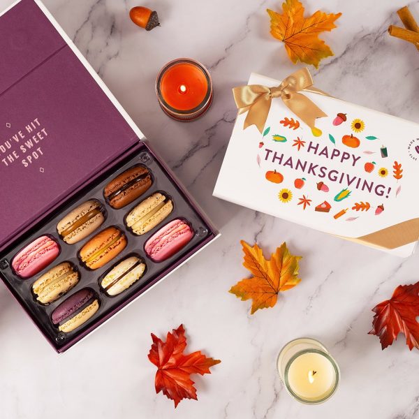 A box full of assorted macarons has a Woops! macaron box with a Thanksgiving sleeve to the right. Surrounding them are some pumpkins, acorns, and paper leaves.