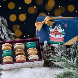 Woops! Christmas macaron gift boxes displayed on snowy decor with frosted pine trees, festive lights, and colorful macarons in teal, brown, and cream tones.