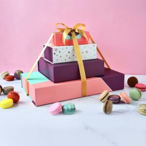 A macaron box of 3, on top of a box of 9, 18, and 27 with a gold ribbon holding them together, and some assorted French macarons lying around