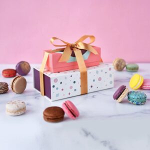 A macaron box of 3 on top of a box of 9 with a gold ribbon holding them together, and some assorted French macarons lying around