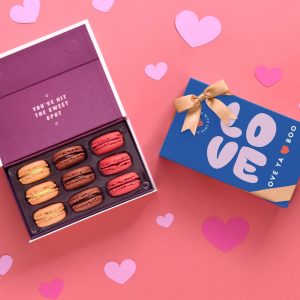 An opened box of 9 French macarons with assorted flavors has a box of 9 French macarons with a Valentine's Day sleeve to its right. Surrouding them are some paper hearts.