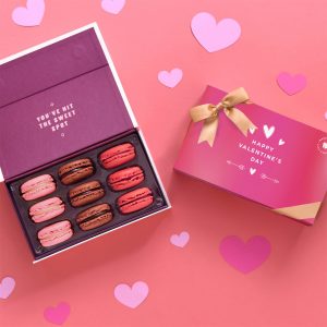 An opened box of 9 French macarons with assorted flavors has a box of 9 French macarons with a Valentine's Day sleeve to its right. Surrounding them are some paper hearts.
