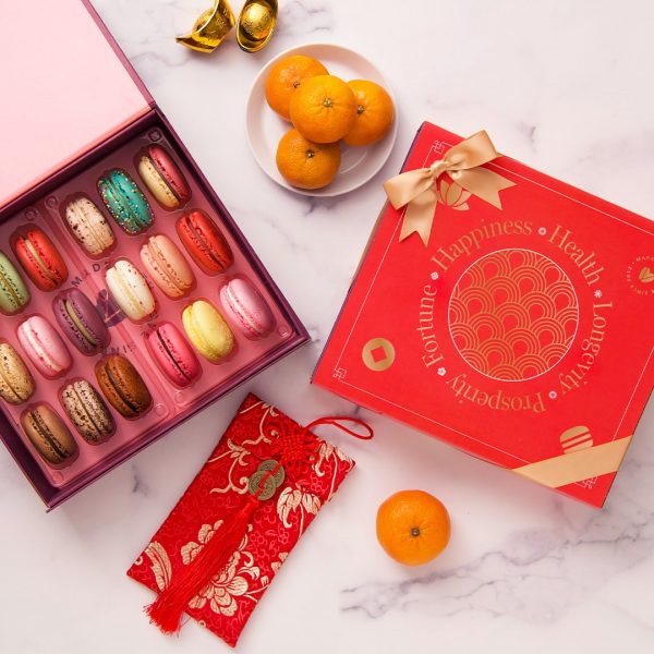 A box full of 18 assorted macarons has a Woops! macaron box with a red Lunar New Year sleeve to the right. Above and below them are some tangerines and Lunar New Year decorations.