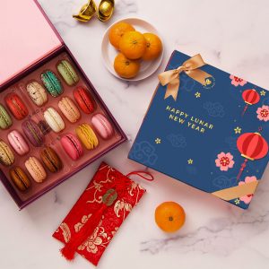 Lunar New Year macaron gift box with assorted colorful macarons, mandarins, and festive red envelope on a marble surface.
