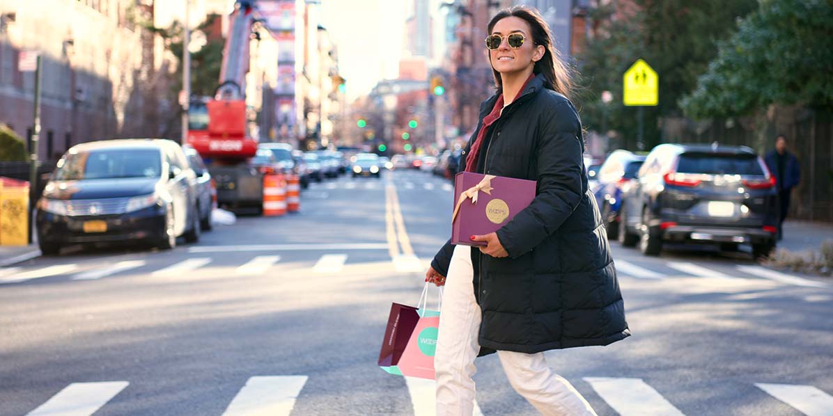 A woman with a black jacket is holding a box and bag of French macarons while crossing the street.