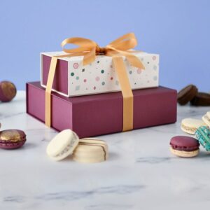 An Argentinian alfajores box of 12 on top of a box of 18 French macarons with a gold ribbon holding them together, and some assorted French macarons and alfajores lying around