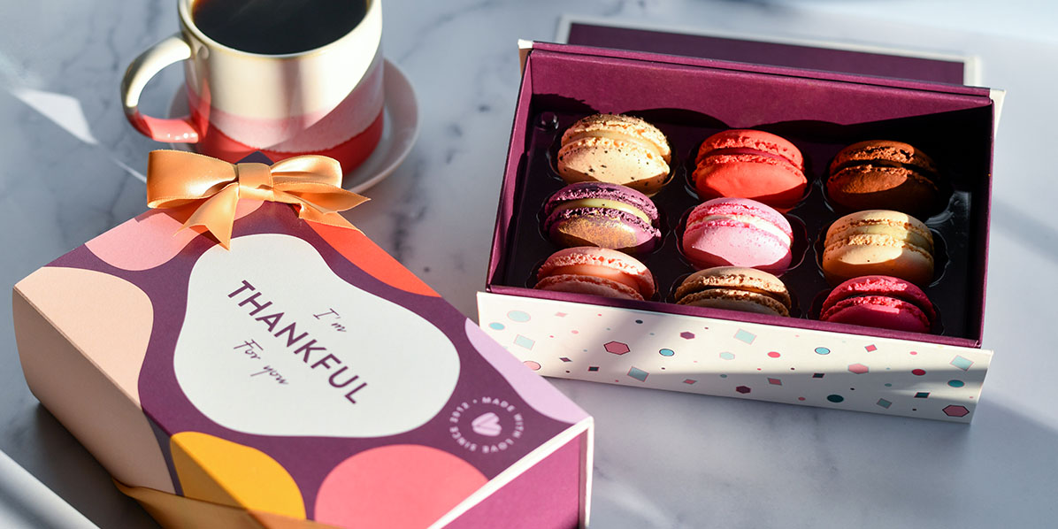 A box of 9 assorted French macarons has a box of French macarons with a Thank You sleeve and a cup of coffee to its left.