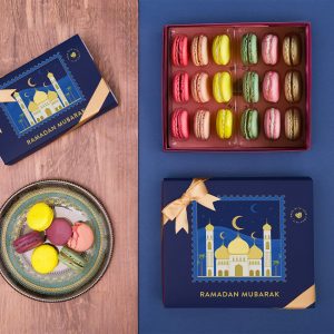 A box full of 18 assorted macarons has two macaron boxes with dark blue Ramadan sleeve to the left and below. On the bottom left corner is a plate full of assorted macarons.