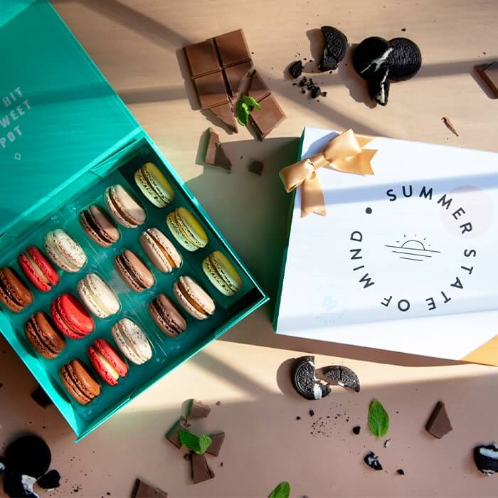 A cream-colored background features a box of 18 French macarons with a “Summer State of Mind” summer sleeve to its right and several chocolates and cookies lying around.