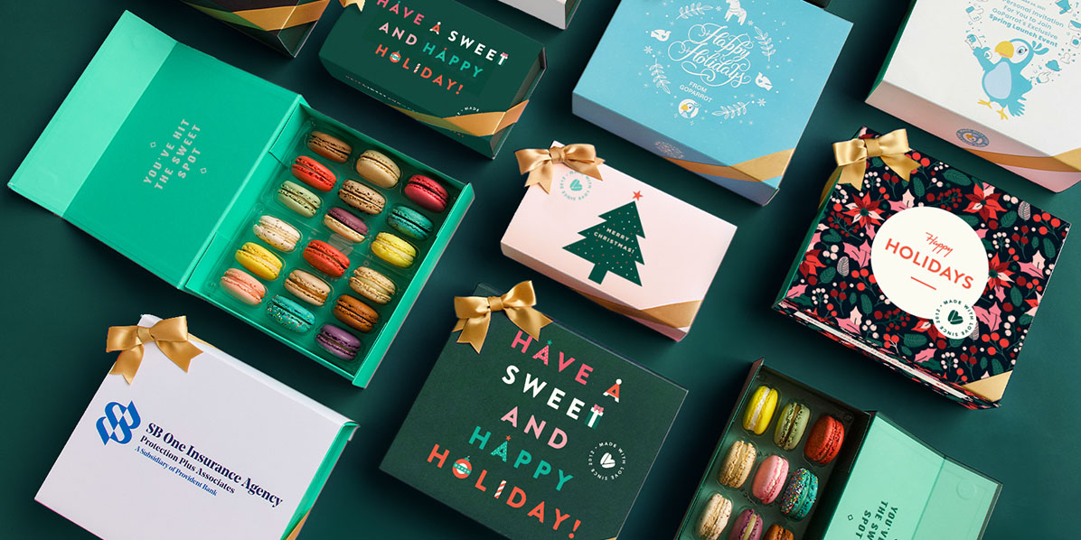 A box full of assorted French macarons is surrounded by several French macaron boxes with Holiday sleeves.