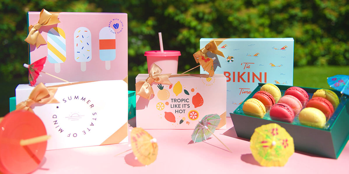 Several French macaron boxes with Summer sleeves are surrounded by cocktail umbrellas, a box full of assorted French macarons, and a pink cup.