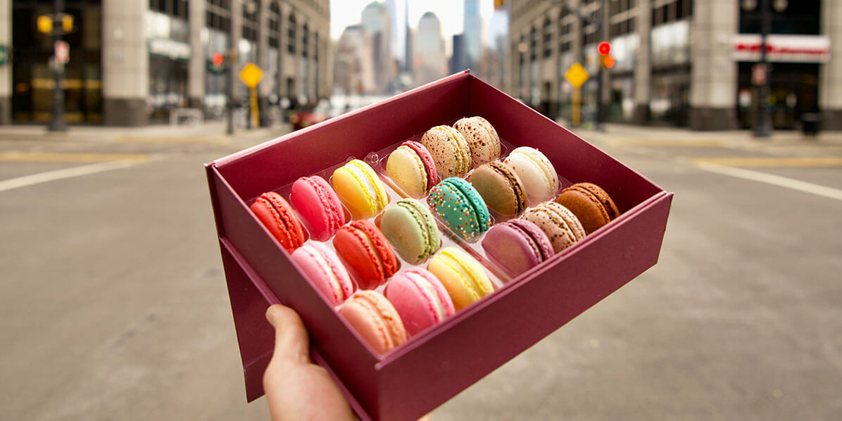 A hand is holding a box full of assorted French macarons in front of a street with buildings.