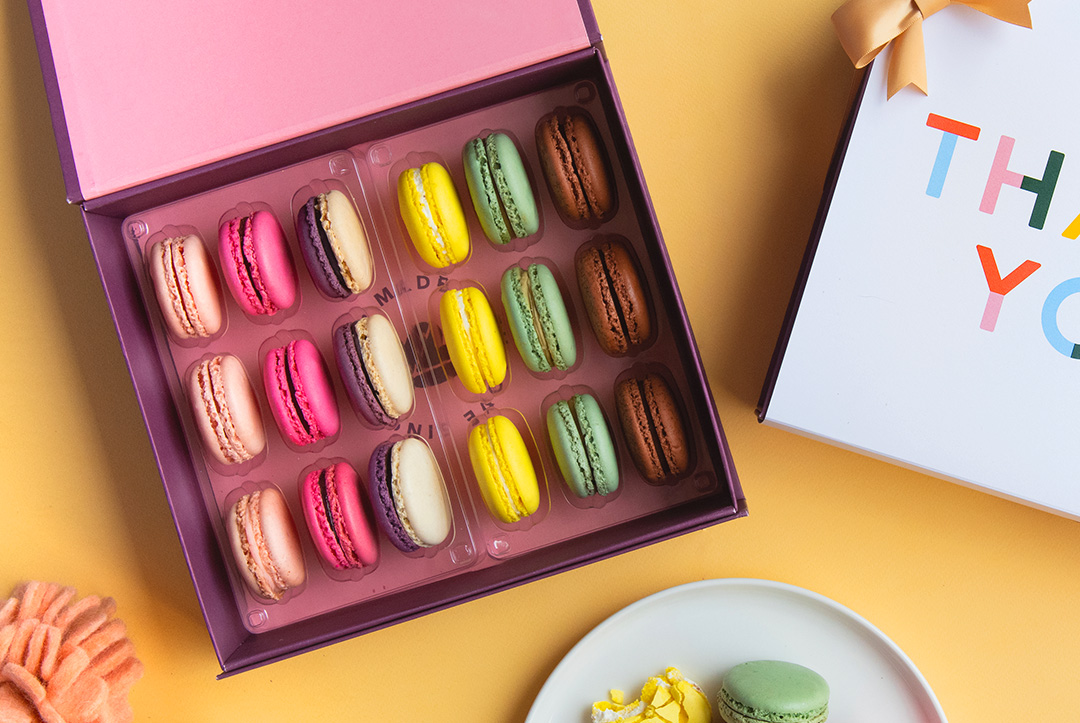 A box full of assorted French macarons has a French macaron box with a “Thank You” sleeve to its right. Below both boxes is a plate full of assorted French macarons.