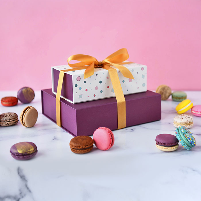 A macaron box of 9 on top of a box of 18 with a gold ribbon holding them together, and some assorted French macarons lying around