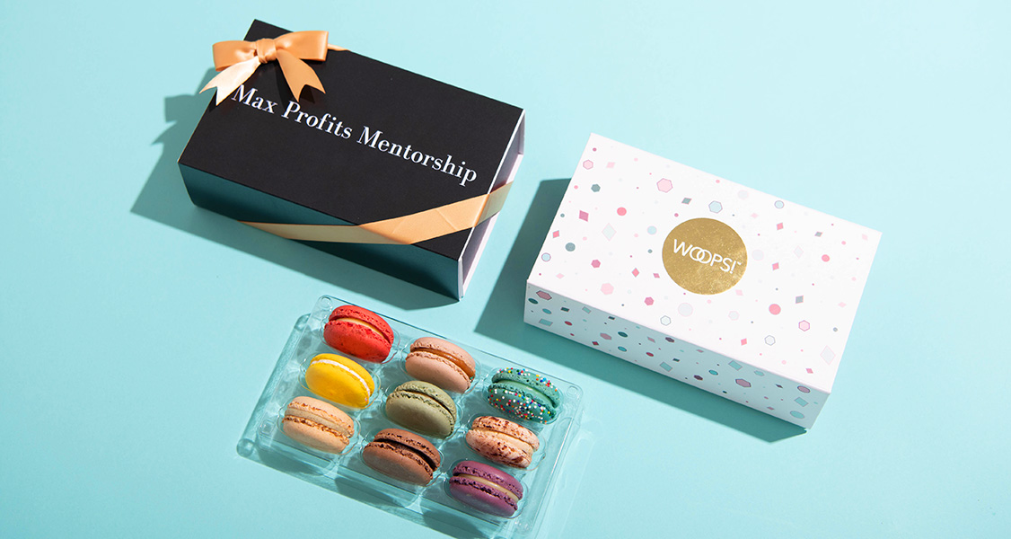 A tray full of 9 assorted French macarons has two French macaron boxes above. One of the boxes has a customized sleeve and a golden ribbon.