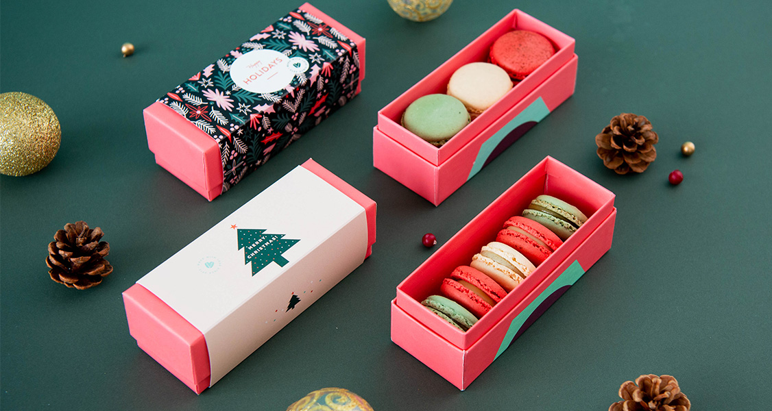 4 French macaron boxes are surrounded by pinecones and holiday ornaments.