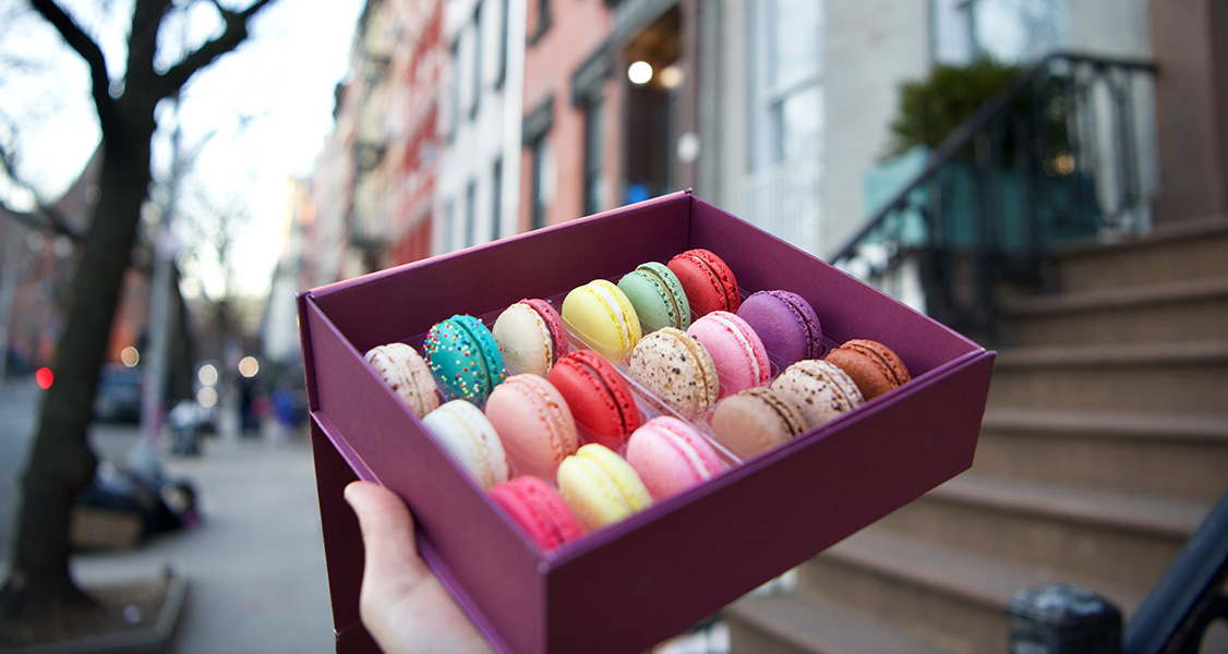 A hand is holding a box full of 18 assorted French macarons in front of a classic New York doorstep.