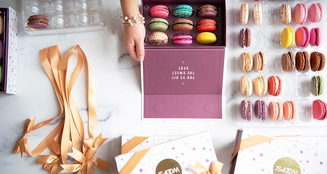 A box full of assorted French macarons is surrounded by trays with assorted French macarons, French macaron boxes, and golden ribbons.