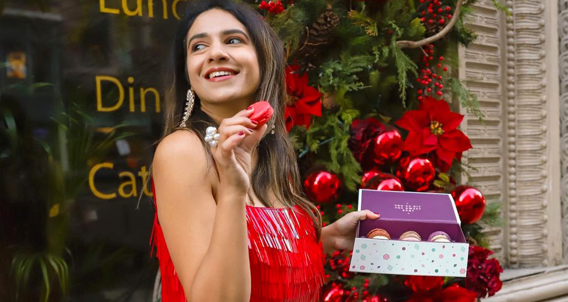 A smiling woman in a red dress is holding a box of French macarons in one hand and a macaron in the other.