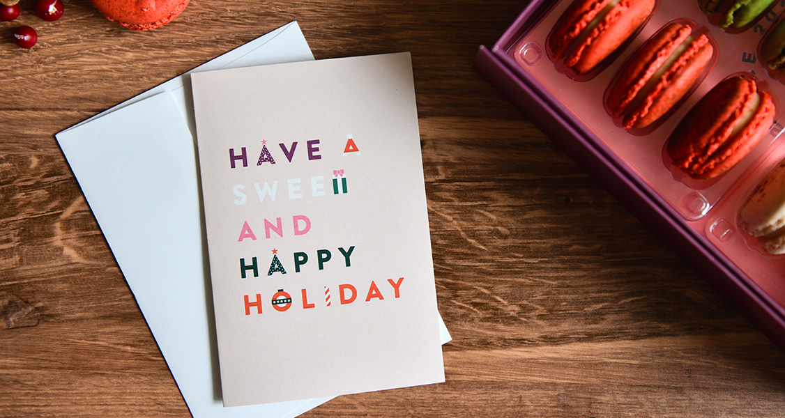 A box with assorted French macarons has a Holiday greeting card to its left. At the top, left corner are two French macarons.